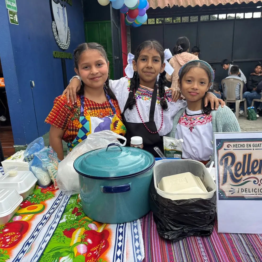 Tres niñas con trajes típicos atendiendo un puesto de comida tradicional en el colegio